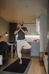 A bearded man practices yoga in his apartment, standing on one leg with his hands folded in front of him meditating