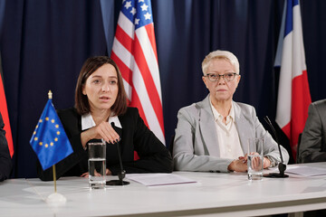 Young female representative of European Union speaking in microphone while sitting in front of...