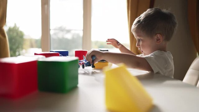 Happy kid is playing at table. Kindergarten education. Boy plays with colored cubes toys. Toddler child builds house from cubes. Boy builder plays with toys at table Preschool education child training