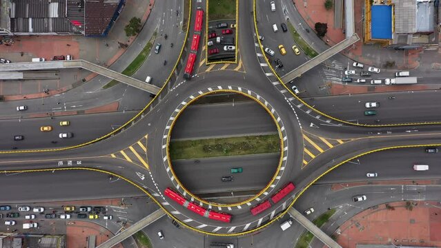 A bird's eye view shot with a 4K drone descending on a round point intersection with red articulated buses, taxis and other vehicles turning from Third street to 30th avenue in Bogotá, Colombia.