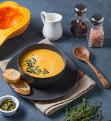 Pumpkin soup puree with cream and seeds in a bowl with bread and fresh herbs on a dark background.