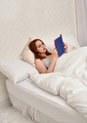 Obraz premium Portrait of beautiful red haired woman wearing comfortable pyjamas, casualy resting in bed, getting ready for sleep, in a glamorous white bedroom location background.