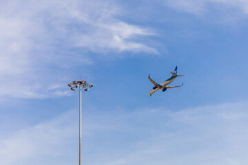 The plane flies next to a pole with searchlights against the blue sky
