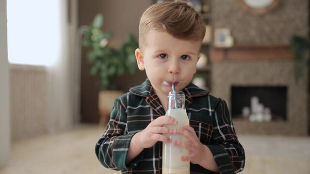 Portrait of cute smiling caucasian toddler boy who drinks milk through a straw. A child in pajamas enjoys drinking yogurt from a glass bottle through an ecological stainless steel drinking straw