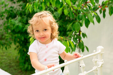 A child covered in strawberry juice stands on a white mettal bed in the garden