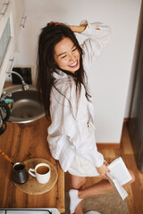 Asian brunette woman in white in kitchen. Female holding, drinking morning cup hot coffee tea - 576633502