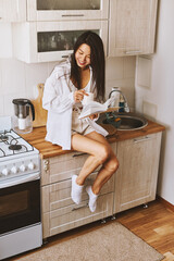 Asian brunette woman in white in kitchen. Female holding, drinking morning cup hot coffee tea - 576633704