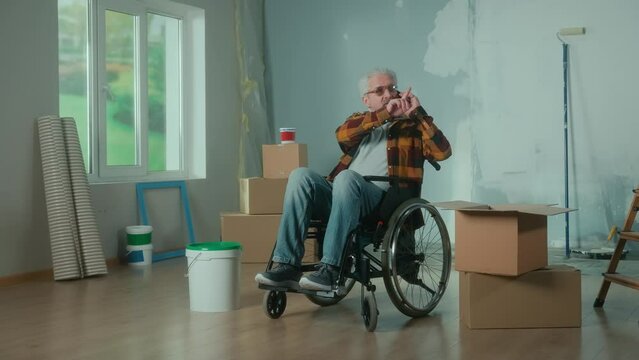An elderly disabled man moves in a wheelchair and talks on a mobile phone. A pensioner takes a roller out of a cardboard box and plans repairs. Room with window, ladder, wallpaper rolls, paint bucket.