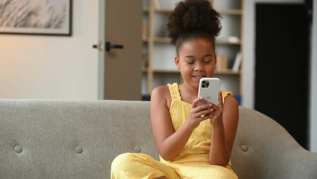 African american girl is sitting on the sofa and using smartphone.