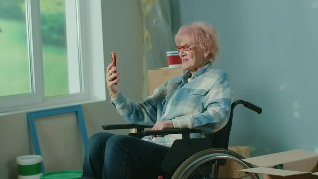 An elderly disabled woman in a wheelchair talks on a video call using a mobile phone. A granny shows a wall and plans repairs. Handicapped person.