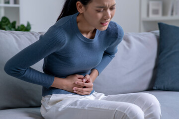 Young Asian woman suffering from strong abdominal pain while sitting on sofa at home. Sick woman...