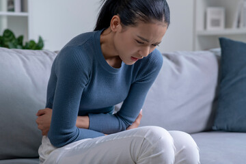 Young Asian woman suffering from strong abdominal pain while sitting on sofa at home. Sick female having a stomach ache.