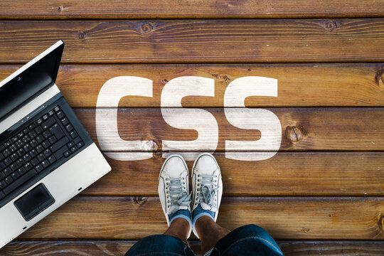 Cascading Style Sheets. Legs in sneakers standing next to laptop and  css on wooden floor
