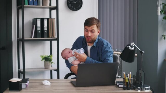 Sitting by table and using laptop. Father with his little baby is at home.