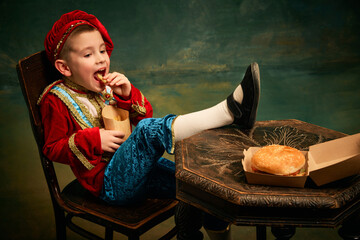 Happy cheerful little boy wearing costume of medieval page boy and prince eating hamburger with...