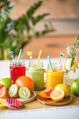 Set of fresh colorful smoothie and juices in three similar glass mugs decorated with straws and bright sliced different fruits on plates on white table in home kitchen. Healthy eating and drinking