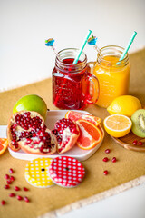 Set of fresh colorful juices in two similar glass mugs decorated with straws and different bright sliced fruits on plates on white table in home kitchen. Healthy eating and drinking
