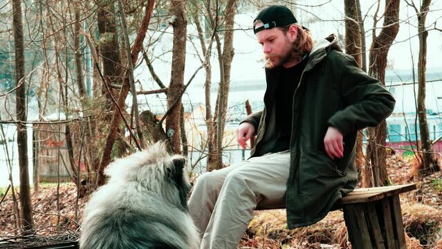 a blond young man in a cap lights a cigarette on a bench while his fluffy friend watches him. The wolfspitz with very bushy fur sits questionably in front of him.