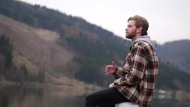 A blond young man throws a stone presumably into a water. After that he puts on a searching funny look and smiles afterwards. In the background you can see a hilly autumn landscape.