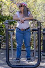 Naklejka premium Senior adult woman standing on a simulator to exercise legs in an outdoor gym, concept of athletic and fitness training in nature, enjoying a sunny day, casual clothes, jeans, hat and sunglasses