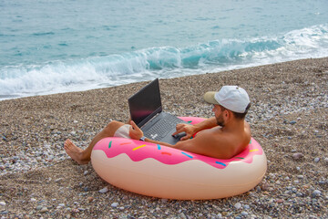Guy in a cap sits in an inflated circle in the form of a donut with pink icing on the beach and works in a laptop online. The concept of employment on vacation, combining work and leisure.