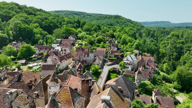 Small, medieval village situated next to a river, flowing through a rich forest in the heart of France. Droneshot flies over the village, showing medieval brick buildings, and walls overgrown by ivy.