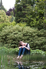 melancholic young woman with long blond hair seating on a bank of a pond in summer park