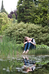 Fototapeta na wymiar melancholic young woman with long blond hair seating on a bank of a pond in summer park