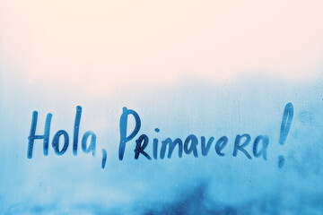 lettering Hola Primavera in Spanish language is Hello spring in english on foggy glass on blue window © ksenija1803z