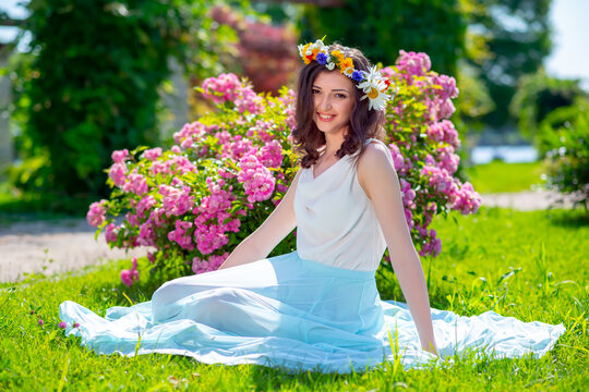 Spring fairy goddess fantasy woman enjoys the sunny day sitting near blooming pink roses. Outdoor close up portrait of young beautiful happy smiling lady. Youth, romantic and lifestyle concept