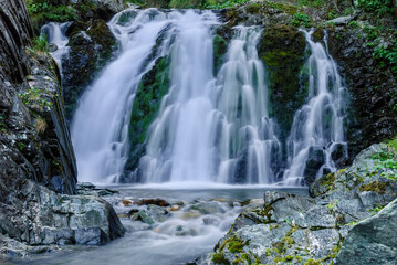 Beautiful mountain rainforest waterfall with fast flowing water and rocks, long exposure. Natural seasonal travel outdoor background with sun shining. Stream waterfall on rocks in the forest