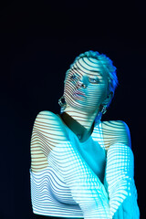Futuristic. Portrait of young blonde girl with neon stripes on face posing over dark background in...