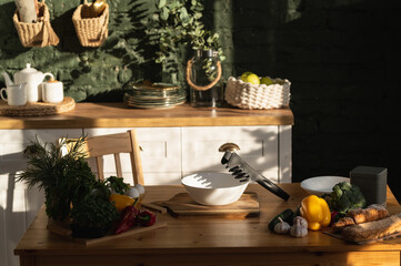 Healthy food kitchen arrangement with salad tongs on a white bowl standing on a wooden board with bell pepper, cucumber, sweet red pepper, broccoli, garlic, croissant, bread, eggs and greens