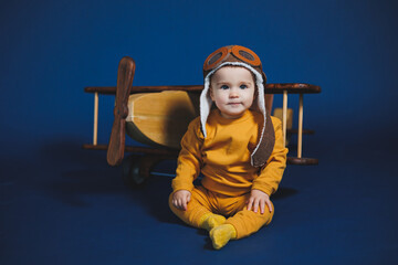 A little boy of 1 year in a yellow knitted suit and an aviator hat stands near a wooden helicopter. Children's wooden ecological toys made of antural wood © DSMT