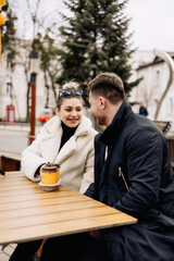 Happy young couple in love in outerwear sitting at a table outside and drinking coffee. Relaxing in the open air