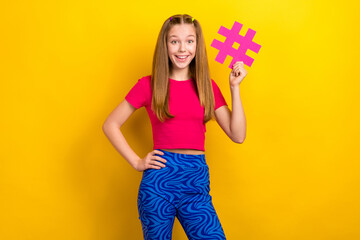 Photo of young excited funny teenager girl wear pink t-shirt crop top touch waist blue retro trousers hashtag isolated on yellow color background