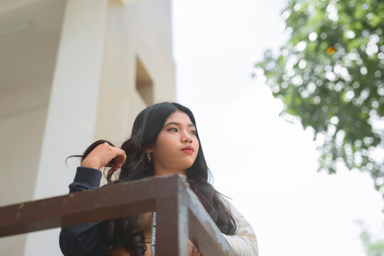 A pensive young asian woman wonders what she did wrong while leaning on the rails of the balcony. Outdoor scene.
