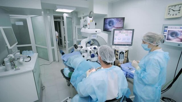 Ophthalmological surgery specialist sits in front of microscope performing operation. Low angle view on the female doctor conducting high-precision surgery. Femto Laser Assisted Cataract Surgery.