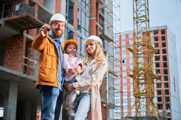 Obraz na płótnie Canvas Man holding apartment keys and smiling while standing next to wife and daughter outside building under construction. Happy family homeowners posing on the street at construction site.