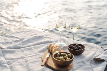 Two glasses of white wine and a wooden plate with bread and olives on a pier at sunset.