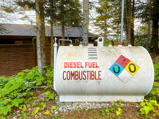 A white diesel fuel storage tank with the words, “Diesel Fuel Combustable” in large lettering on the front. Colorful environment warning symbols are painted on the tank. Wilderness in background.