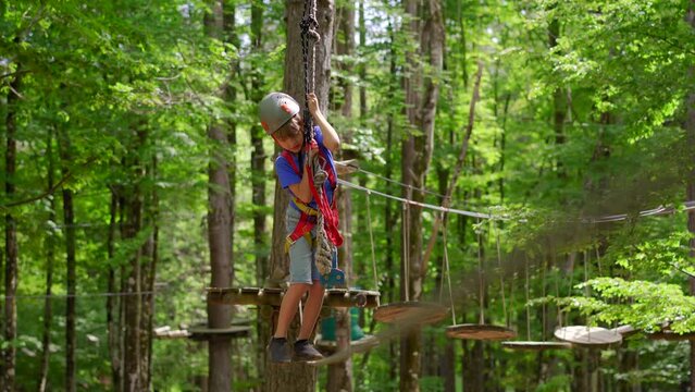 A little boy is enjoying an exciting adventure on a rope obstacle course in a lush forest. He is wearing a helmet and a safety harness as he navigates through the challenging course, showing bravery