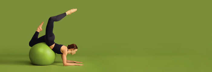Young slim fit girl doing stretching exercises with fitness ball over green studio background....
