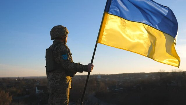 Soldier of ukrainian army holding waving flag of Ukraine against background sunset. Man in military uniform and helmet lifted up flag. Victory against russian aggression. Invasion resistance concept