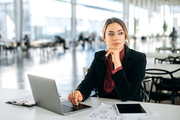 Photo of a pensive thoughtful elegant caucasian business woman, executive, recruitment, product manager, sits in a business center, working in a laptop, tired looking away, thinking, dreaming