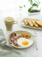 Plate of fried egg, bacon, baked beans, fresh toasts and cup of coffee on white marble table in front of window. English breakast minimal style