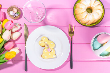 Cute Easter table setting, spring holiday decorated table with plate, glass, cutlery, tulip flowers bouquet, Easter decoration symbols and treats, on pink wood bright table, top view copy space