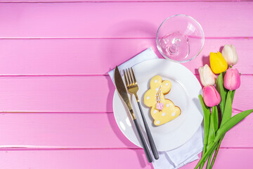 Cute Easter table setting, spring holiday decorated table with plate, glass, cutlery, tulip flowers bouquet, Easter decoration symbols and treats, on pink wood bright table, top view copy space