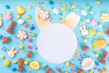 Easter sweets and candies background. High-colored blue flat lay with various Easter chocolate eggs, treats, symbols of easter sheep, bunny, carrot, flowers, cookies, top view copy space, frame
