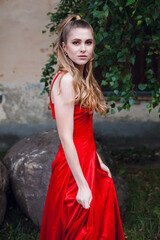 A cute young girl with blond flowing long hair and beautiful make-up, in a long red dress, stands on the street of the old city. There is a tree nearby.